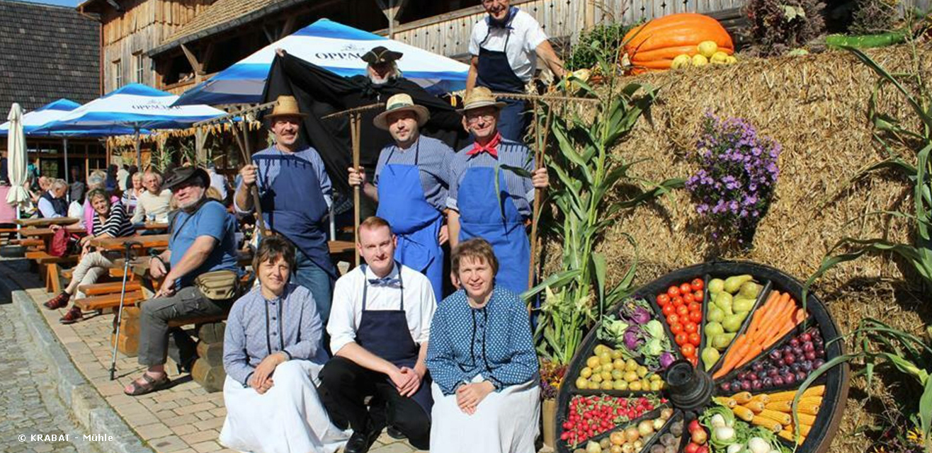 Eine Tanzgruppe sitzt beim Erntedankfest in der Krabat – Mühle in Schwarzkollm vor bunten Marktständen.