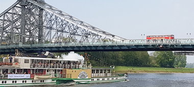 Ein Dampfschiff fährt auf der Elbe unter dem Blauen Wunder lang und ein Roter Doppeldecker überquert die Brücke.